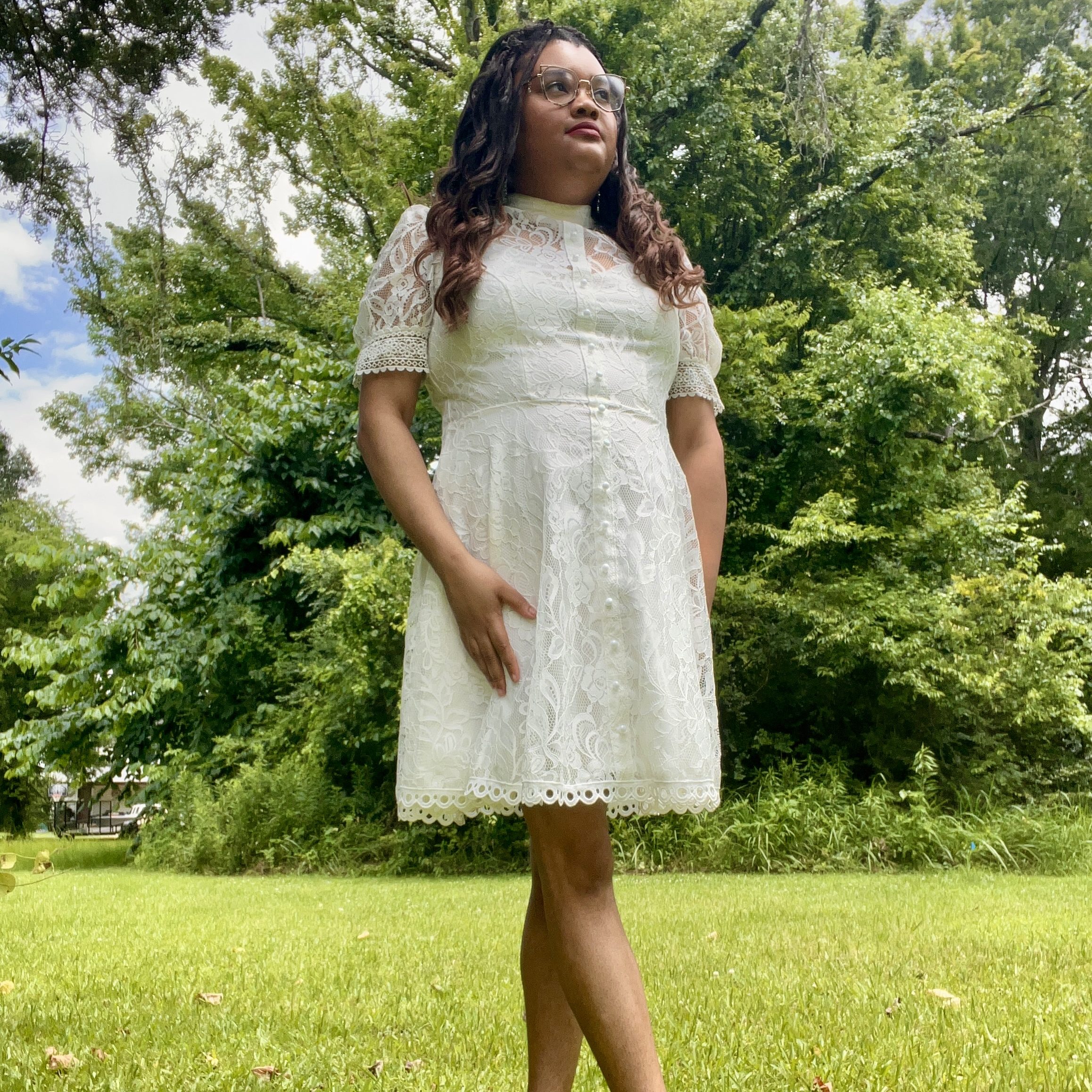 Woman in a white lace dress and white heels standing on green grass, surrounded by lush trees under a partly cloudy sky.